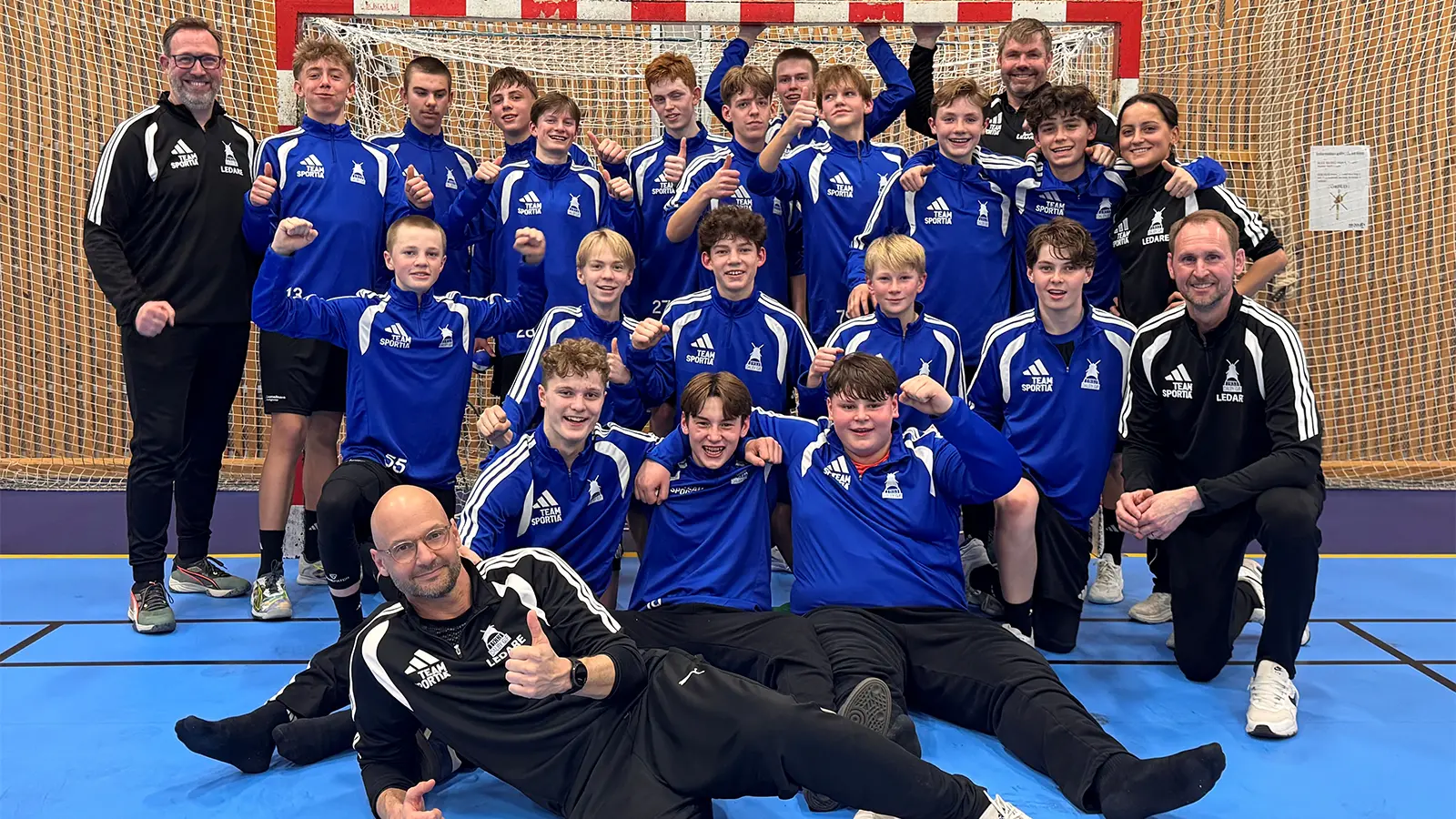 Ett lag med killar och tränare står och sitter ihop framför ett handbollsmål